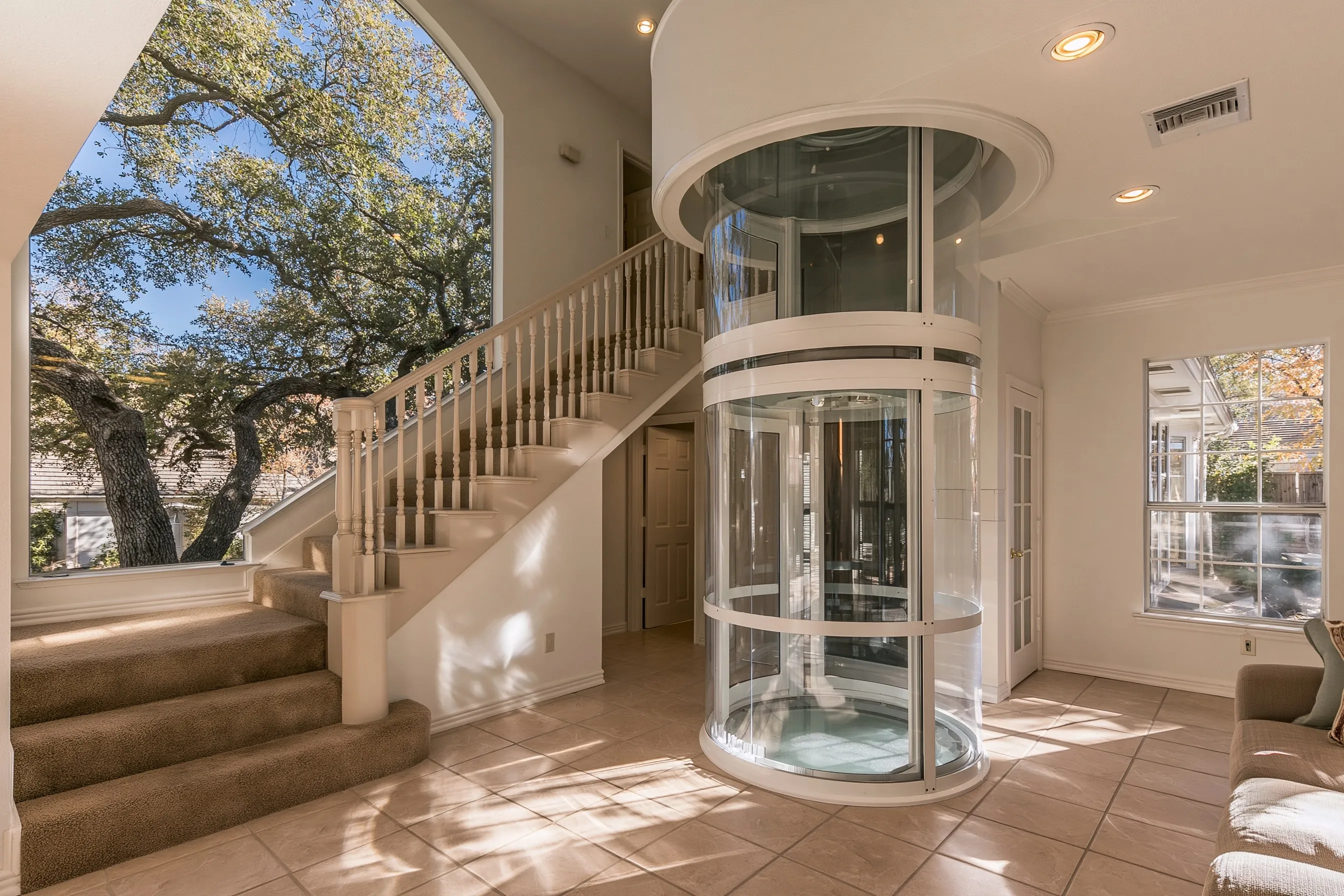 Wide round clear vacuum elevator on a tile floor beside a carpeted stair, rising into a round ring in the ceiling, a live oak through the tall window
