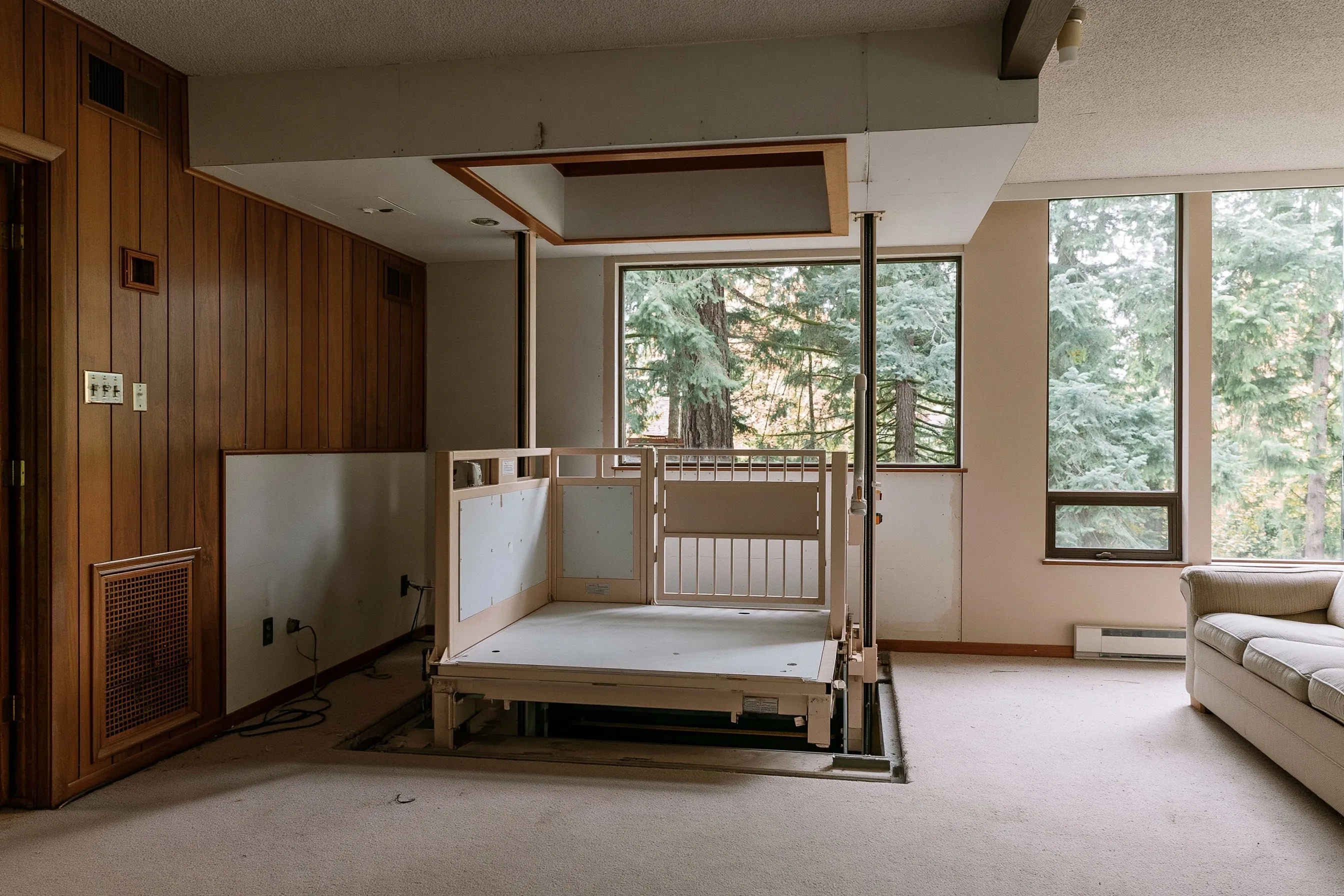 Wheelchair-size platform lift with side panels and a gate in a wood-paneled family room, below a framed opening in the ceiling