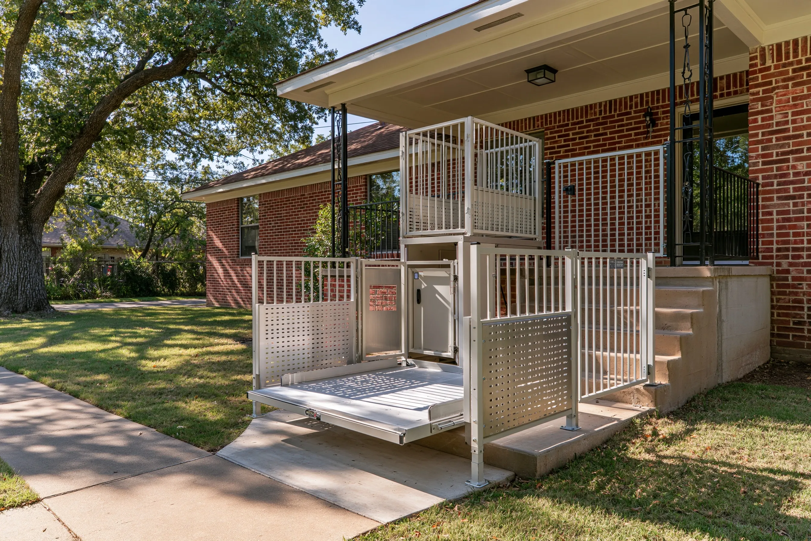 Vertical wheelchair platform lift with guard panels and a gate beside the front porch steps of a single-story brick house